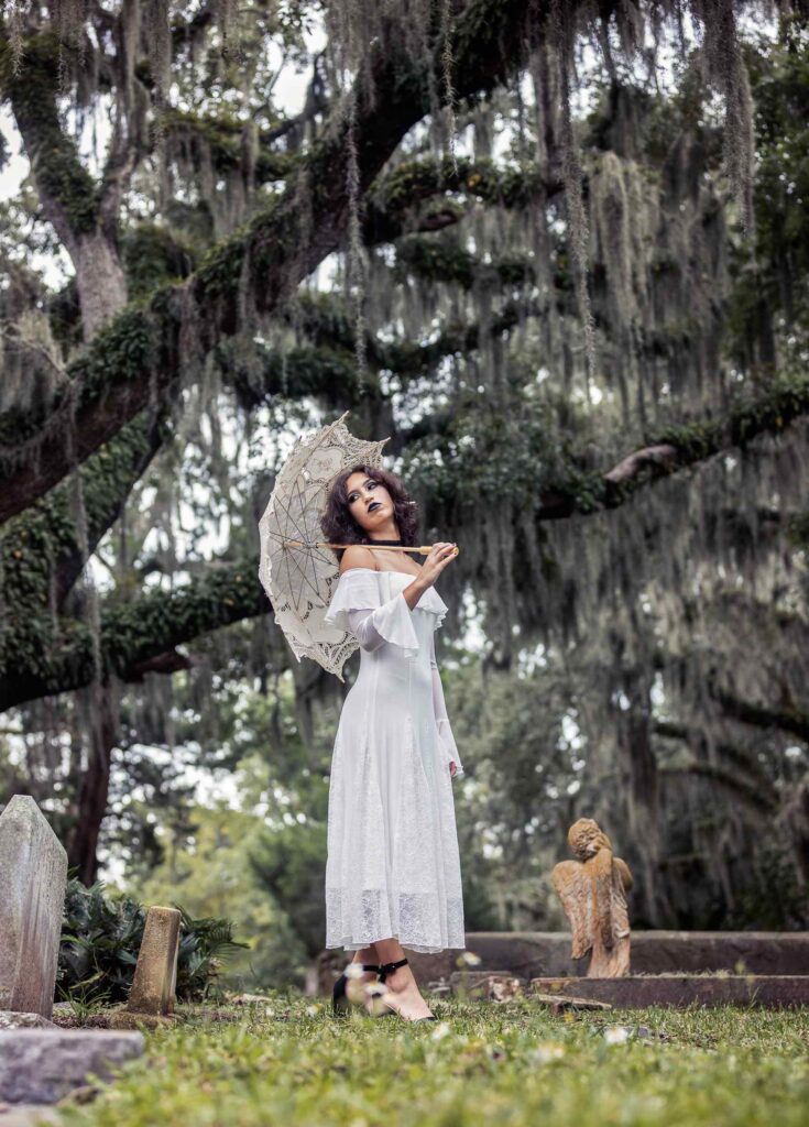 North Florida senior portrait of a high school senior standing beneath dramatic Spanish moss with a lace parasol in an Amelia Island cemetery, styled with a soft gothic aesthetic and photographed by Nature’s Child Photography, a Jacksonville senior photographer